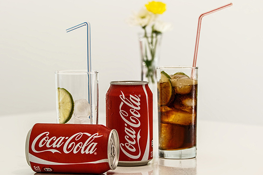 Coca Cola cans next to a glass of ice and a glass of Coca Cola.